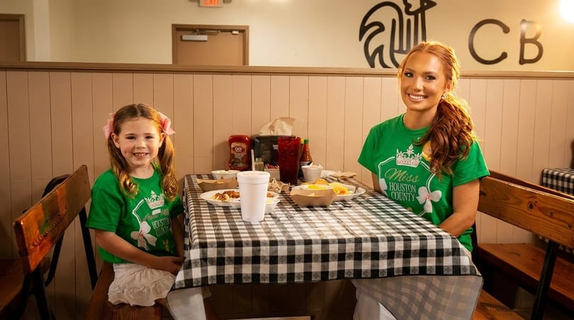 Two smiling females, a woman and a young girl, wearing green shirts, seated at a checkered table with food.