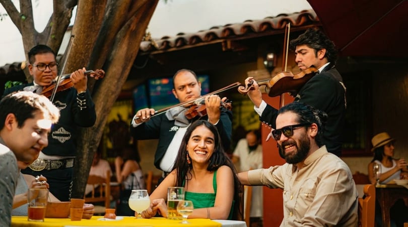 Mariachi band playing violins for smiling guests enjoying drinks at an outdoor restaurant.