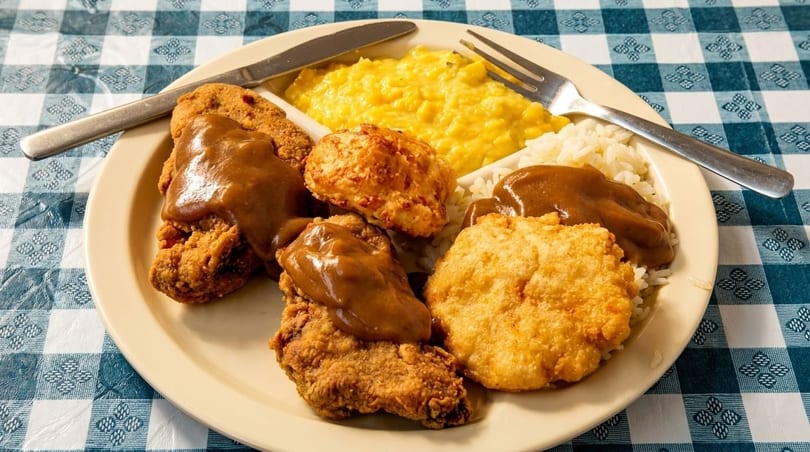 A plate of fried chicken with gravy, rice, corn, and biscuits on a blue and white checkered tablecloth.